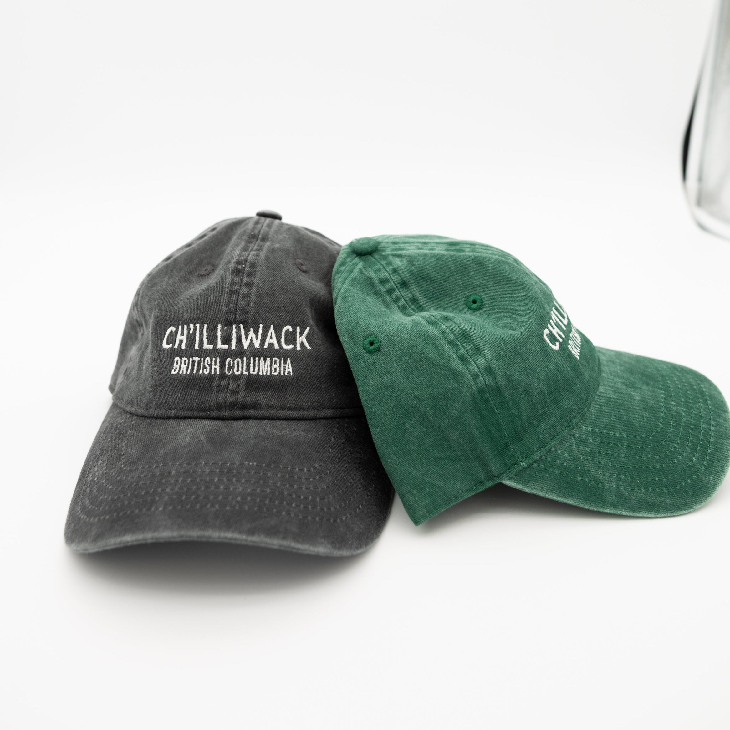 Two baseball caps on a white background: one dark gray cap embroidered with 'CHILLIWACK BRITISH COLUMBIA' in white, and one green cap with white embroidered lettering on the side.